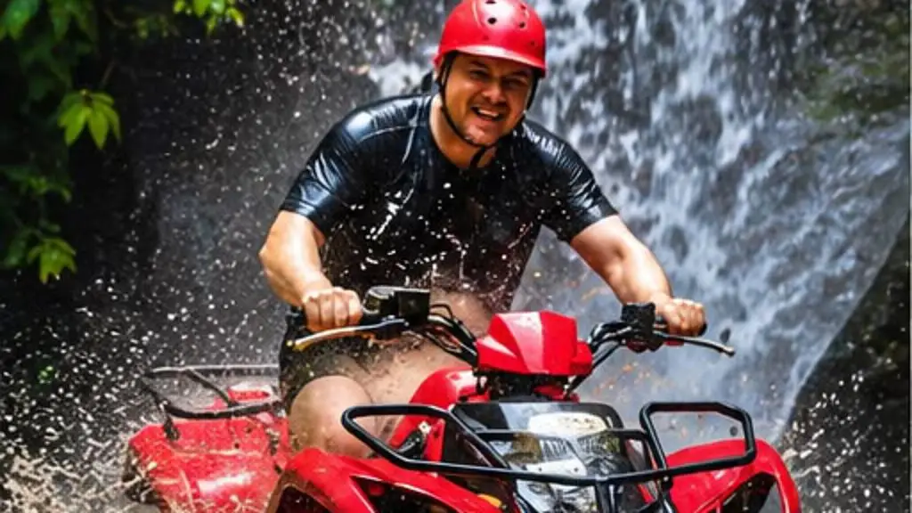 A smiling rider in a red helmet on a red quad bike, splashing through shallow water beneath a waterfall