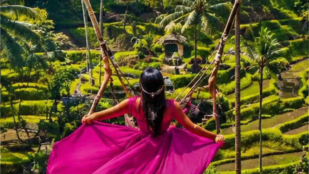 A woman in a long pink dress on a rope swing above green rice terraces and palm trees, with the Alas Harum sign on the hillside