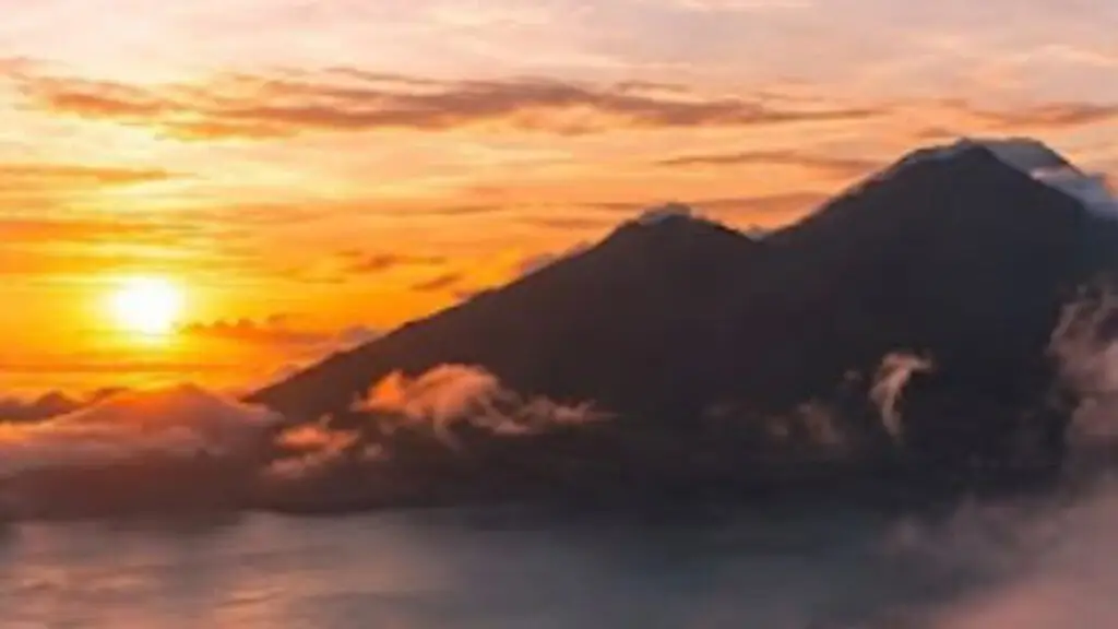 The sun rising orange beside the dark cone of Mount Batur, with a layer of cloud filling the valley below