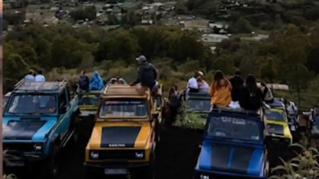A row of blue and yellow open-top jeeps parked on a ridge at sunrise, with travellers standing on them looking over the caldera and lake