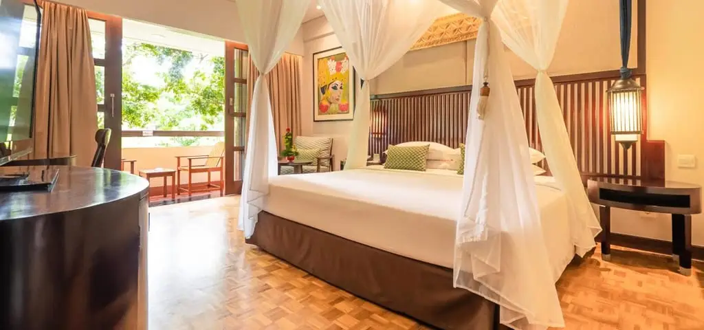 A resort bedroom with a white four-poster canopy bed on a parquet floor, a carved timber headboard, and doors open onto a balcony with trees beyond