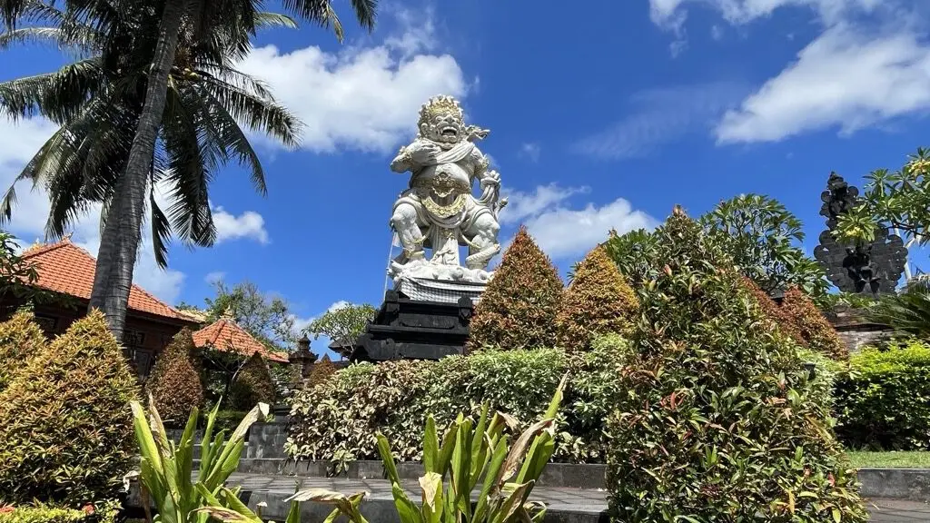 A carved Balinese guardian statue standing in a trimmed hotel garden of palms and clipped shrubs under a blue sky