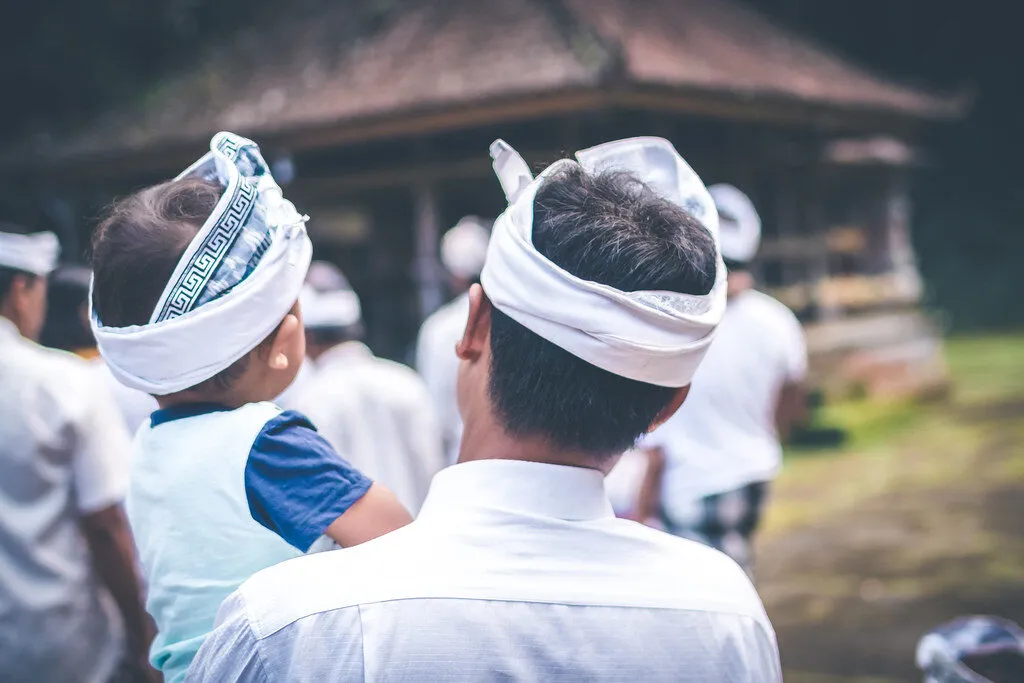 A Balinese man in a white udeng headband carrying a child on his hip in a temple courtyard