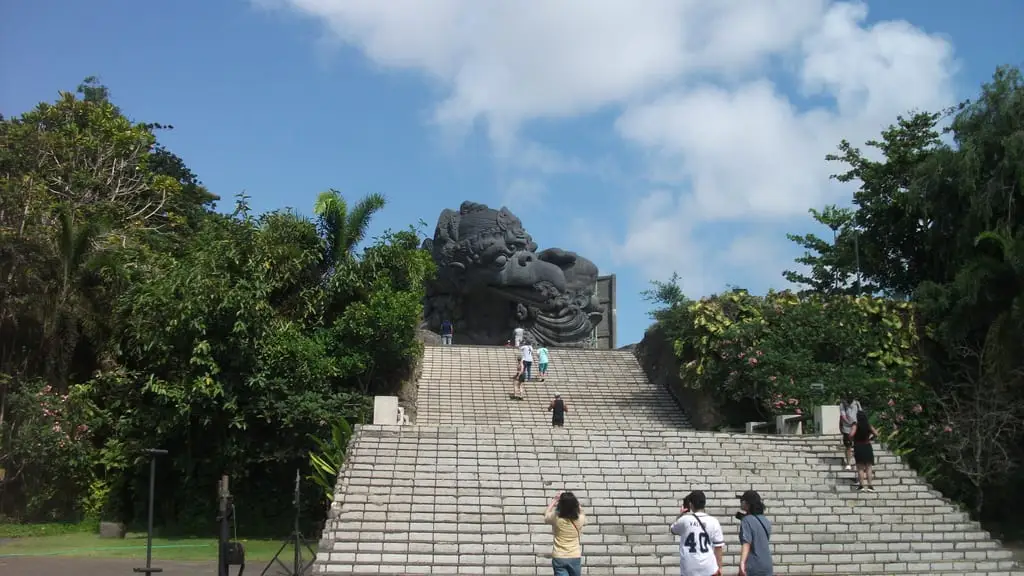 Wide stone steps climbing to the dark head of the Garuda Wisnu Kencana statue under a blue sky with white clouds