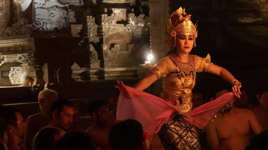 A dancer in a gold costume and tall crown, arms spread, performing before a seated audience in a torch-lit stone courtyard