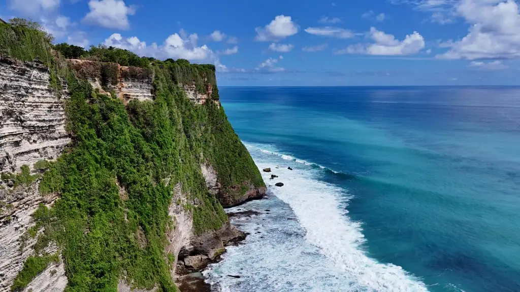 Limestone cliffs covered in green scrub dropping into deep blue sea with white surf, under a clear sky