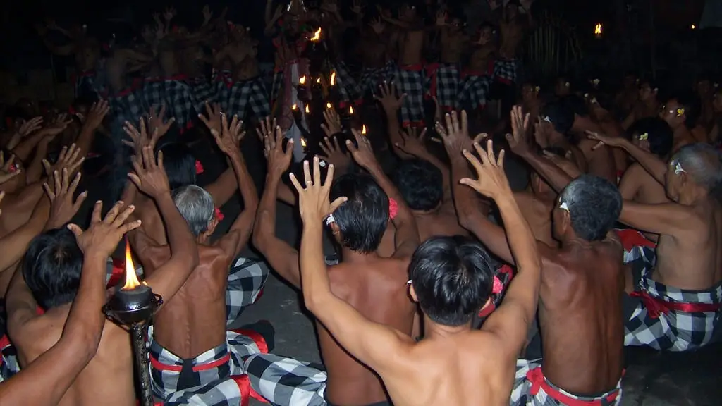 A ring of bare-chested Kecak dancers in checked sarongs raising their arms around a flaming torch at night