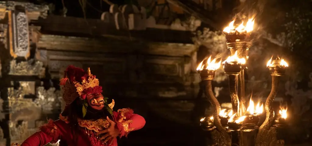 A Kecak performer in a red costume and gold headdress beside a torch stand of open flames in a stone courtyard at night