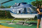 A couple embracing beside a blue and white helicopter on a green and yellow helipad, with the sea and a forested headland behind them