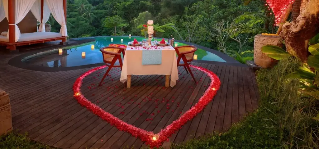 A dinner table for two on a timber deck at dusk, ringed by a heart of red rose petals, beside a candlelit private pool and a curtained daybed pavilion, with the jungle behind