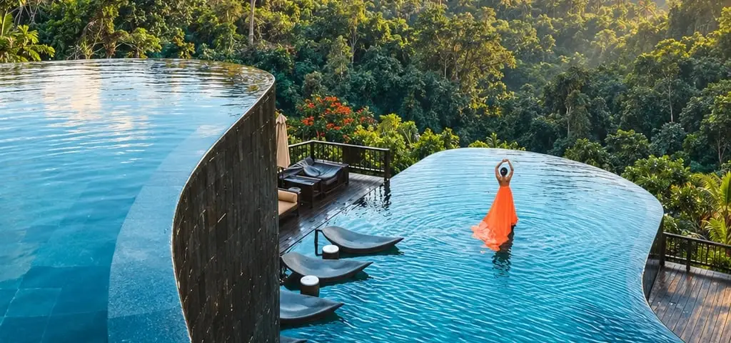 Two infinity pools on different levels curving out over a jungle valley, a woman in a long orange dress standing in the lower one with her arms raised, loungers set into the shallows beside a timber deck